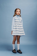 Full length studio portrait of girl in cream apron dress with blue floral stripe print, light blue turtleneck, white socks and brown shoes, direct gaze