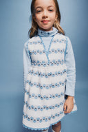 Close up of cream apron dress with blue floral stripe embroidery and scalloped trim, worn over a light blue turtleneck with embroidered collar