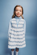 Close up studio portrait of girl in cream apron dress with blue floral stripe print and light blue turtleneck, smiling