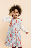 Toddler girl wearing blue and white striped pinafore dress with red rose print, cream long sleeve top underneath, covering one eye playfully