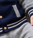 Close up of navy cashmere letterman jacket hem with striped ribbed trim, wooden buttons, and patch pocket, worn with grey leggings