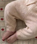 Close up of baby's legs in cream pointelle knit pants with pink scalloped cuff trim, lying on apple print blanket