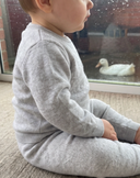 Side view of baby sitting in light grey cashmere sweater and pants set by a rain streaked window, duck visible outside