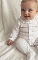 Baby wearing white pyjama sleepsuit with ruffled collar and gold embroidered trim, sitting on white blanket