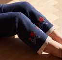 Close up of bent knees in navy knit pants with red rose embroidery and cream ribbed cuffs, on wood floor