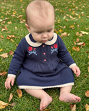 Baby sitting cross legged in navy knit dress with cream collar and red rose embroidery, on grass with fall leaves