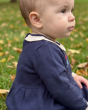Side profile close up of baby in navy knit dress with cream collar and red rose embroidery, on grass