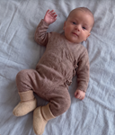 Baby wearing brown knit wrap romper and cream wool booties, smiling on striped sheet