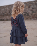 Full length back view of girl in navy dress with red stitched bow, tiered ruffle skirt, standing barefoot on beach near stone wall