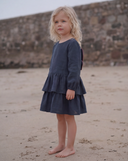 Girl wearing navy dress with tiered ruffle skirt, standing sideways on sandy beach, looking off camera