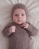 Close up portrait of baby wearing brown knit bonnet, hands clasped near chest, wrap romper with tie closure