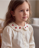 Toddler girl wearing cream lace blouse with red rose embroidered collar, looking to the side while seated