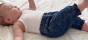 A baby lying on a white bed, wearing a white ribbed tank top and blue denim shorts.