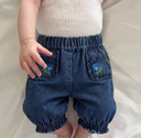 Close up of baby standing in dark wash denim harem pants with floral embroidered pockets
