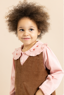 A young child with curly brown hair wearing a pink long-sleeve shirt with a ruffled Peter Pan collar and a brown corduroy pinafore-style vest, photographed against a plain warm beige background.