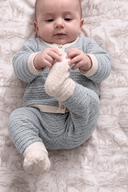 A baby lying on a patterned quilted blanket, wearing a light blue-gray ribbed knit one-piece with cream trim and matching cream knit socks.