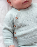 A close-up of a baby wearing a pale sage green knit cardigan with white leaf patterns and brown buttons, lying on a white fabric surface.
