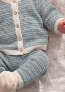 Close-up of a baby wearing a light blue and white striped knit cardigan and matching booties, lying on a patterned white sheet.