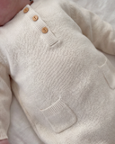 Close-up of a cream-colored knit baby romper with a three-button henley neckline and a front patch pocket, shown on a baby lying on a soft white blanket.