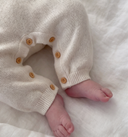 Close-up of a baby's bare feet wearing cream-colored knit pants with small wooden buttons, lying on a soft white textured blanket.