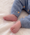 Close-up of a baby's bare feet resting on a soft, textured off-white blanket, wearing a light blue cashmere romper with small wooden buttons.
