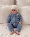 A baby sitting on a white fabric surface, wearing a light blue cashmere romper matching cream knit hat.