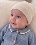 A close-up of a baby wearing a cashmere beanie and a blue cashmere romper with a white collar, resting against a soft cream pillow.
