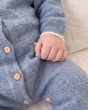 Close-up of a baby wearing a light blue cashmere romper with wooden buttons and a cream ribbed cuff, resting on a soft beige surface.