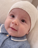 A close-up of a baby wearing a cream-colored cashmere beanie and a blue cashmere romper with a peter pan collar