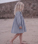 A young child with long blonde hair walks barefoot on a sandy beach, wearing a blue and white gingham plaid long-sleeve dress.