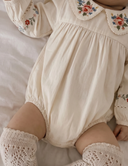 Close up of baby wearing ecru bubble romper with floral embroidered collar and lace trim ankle socks, lying on white bedding