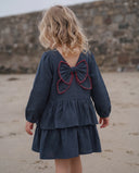 Girl wearing navy dress with red stitched bow at back, tiered ruffle skirt, walking away on sandy beach in front of stone wall