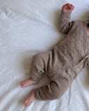 Baby lying on white sheet wearing taupe pointelle knit kimono jumpsuit, full body flat lay lifestyle shot with bare feet