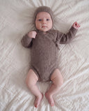 Baby lying on bed wearing brown melange knit wrap romper and matching bonnet, both hands raised in fists