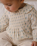A close-up of a baby wearing a cream-colored long-sleeve smocked romper with a ruffled Peter Pan collar and a blue and green floral print, paired with matching cream ribbed pants.