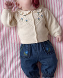 A baby lying on pink and white striped bedding, wearing a cream-colored knitted cardigan with blue floral embroidery and dark blue denim pants with matching embroidered pockets.