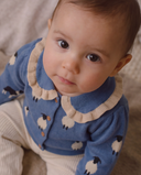 A close-up of a baby wearing a blue long-sleeve cardigan with a cream ruffled collar and sheep pattern, paired with cream ribbed pants.