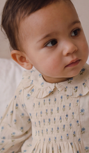 A close-up of a baby wearing a cream-colored smocked dress with a ruffled Peter Pan collar and small blue floral prints.