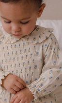 A close-up of a young child wearing a cream-colored smocked dress with a blue and green floral pattern, featuring a ruffled collar and long sleeves.