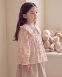 A young girl with long dark brown hair stands in profile wearing a cream-colored long-sleeve blouse with a pink floral pattern and a ruffled Peter Pan collar, paired with a beige skirt. She is posed in a softly lit room with plush teddy bears visible in the background.