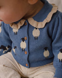 A close-up of a baby wearing a blue knit cardigan with a cream ruffled collar and knitted sheep pattern, paired with cream ribbed pants.