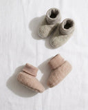 Two pairs of soft knit baby booties in neutral tones, one pair in light taupe and one pair in dusty blush, arranged on a white textured fabric background.