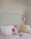 A baby in cream patterned pajamas lies on a bed with white bedding, a light sage green upholstered headboard, and a striped pillow, against a neutral beige wall with a wall-mounted lamp.