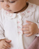 Close up of baby wearing white sleepsuit with scalloped collar and pink floral embroidery, hand resting on chest