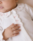 Extreme close up of baby wearing white sleepsuit with scalloped collar and pink floral embroidery, hand touching collar