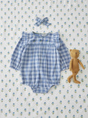 A baby girl's long-sleeve smocked bodysuit in a blue and white gingham check, styled in a flat lay with a matching blue and white gingham bow headband and a small brown teddy bear on a white muslin fabric background with a blue floral print.