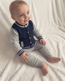 Baby boy wearing navy and grey letterman cardigan with striped cuffs and matching leggings, sitting on white fur blanket