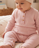 Baby sitting in light pink cashmere romper with wooden buttons and patch pockets, wearing a matching cream knit hat, on cherry print bedding