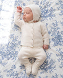 Newborn wearing off white cashmere three piece set, jeweled button cardigan, footed trousers, and matching bonnet, lying on blue floral bedding