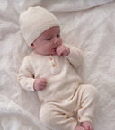 A baby lying on white textured bedding, wearing a cream cashmere one-piece romper with a buttoned cashmere matching beanie.